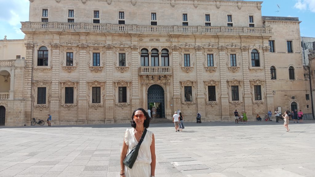 Author Lorena Basualdo standing in Piazza del Duomo in Lecce, Puglia, with the cathedral, bell tower, and municipal buildings in southern Italy.